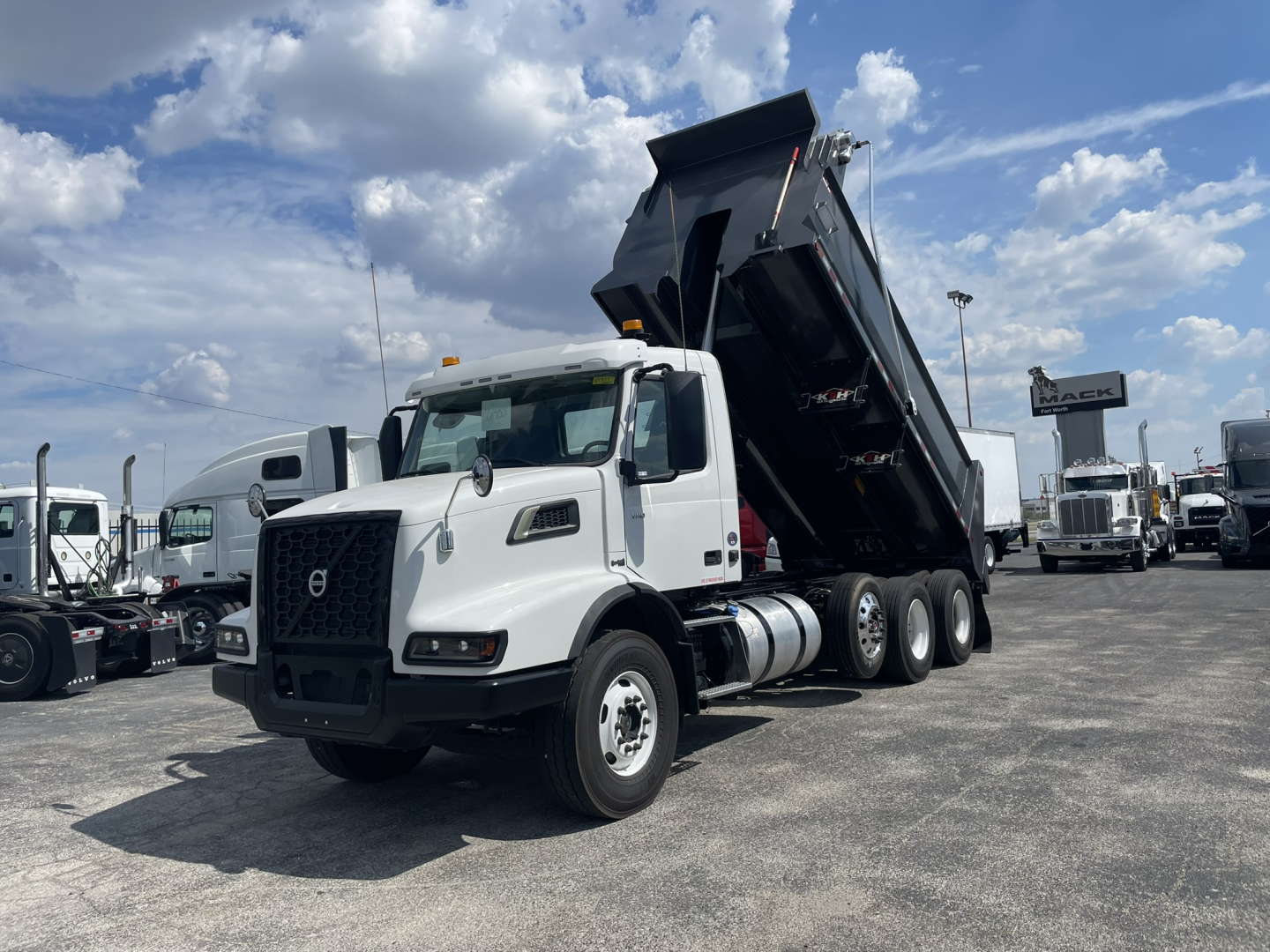 Volvo VHD86B300 dump truck in a dealership lot, white cab with black dump bed raised, multiple trucks in background.