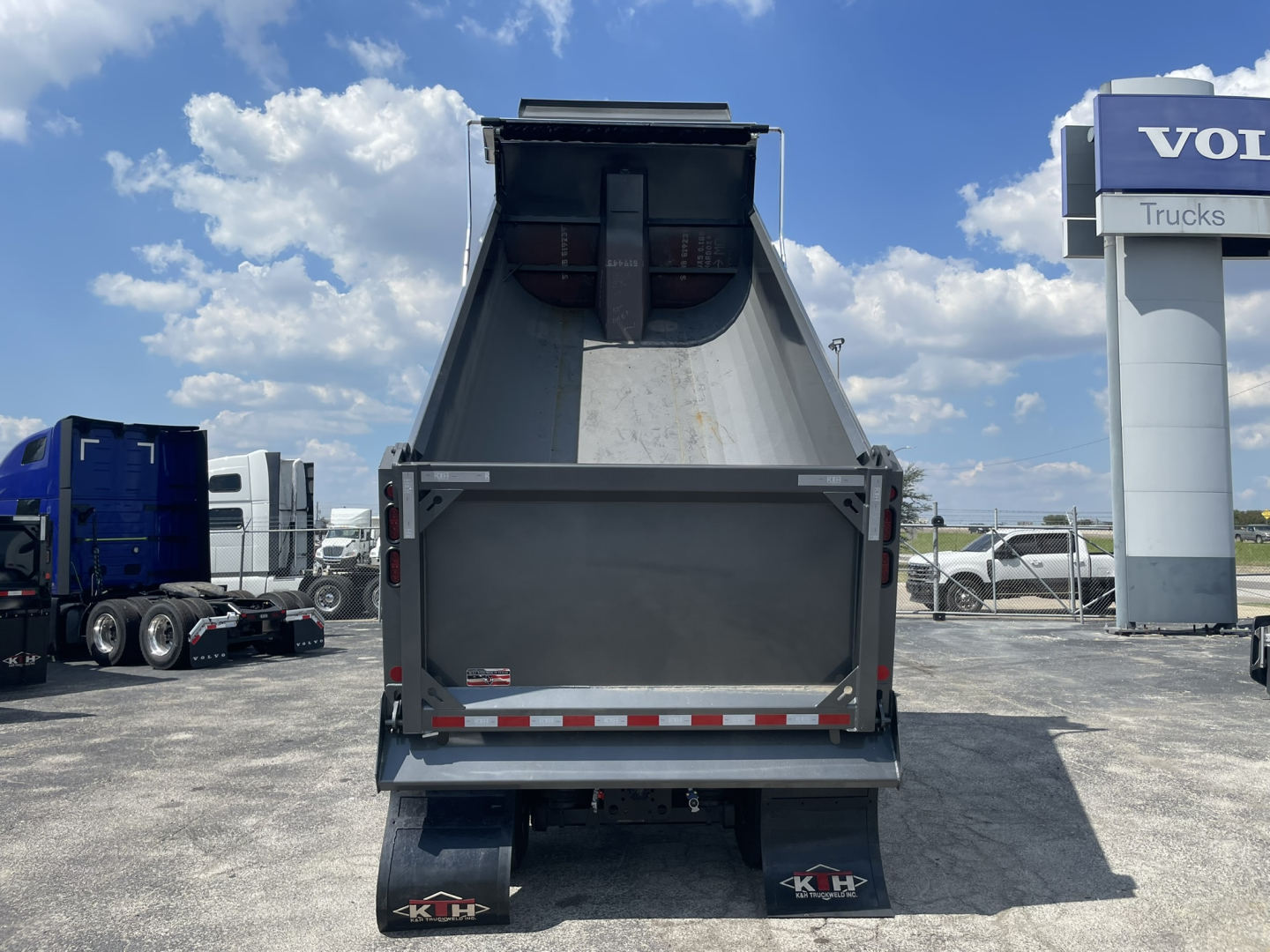 Rear view of a Volvo VHD86B300 dump truck bed in a dealership lot. View shows the open-top dump body with tailgate, rear lights, and mudflaps. Visible signs include a Volvo Trucks sign in the background and other trucks parked nearby.