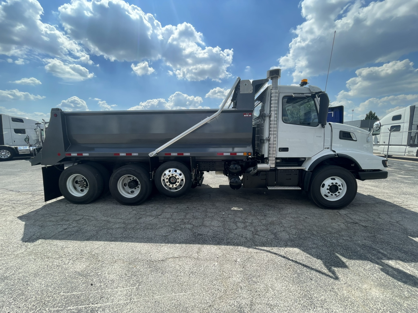 Volvo VHD86B300 dump truck side view showing six wheels, white cab, gray dump bed, hydraulic lift, and safety rails.