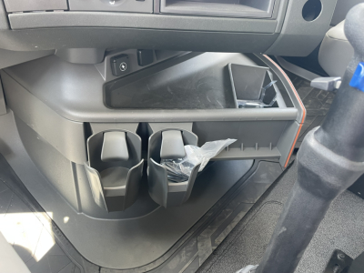 Center console area of a Volvo VHD86B300 cabin showing cup holders, small storage compartments, and a partially open lower cubby with a plastic bag inside.