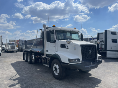 Volvo VHD86B300 dump truck in a dealership lot, white cab with black grille, chrome mirrors, and dual rear axles. Visible text includes Volvo emblem on grille and generic dealership context.