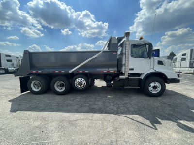 Volvo VHD86B300 dump truck side view showing six wheels, white cab, gray dump bed, hydraulic lift, and safety rails.