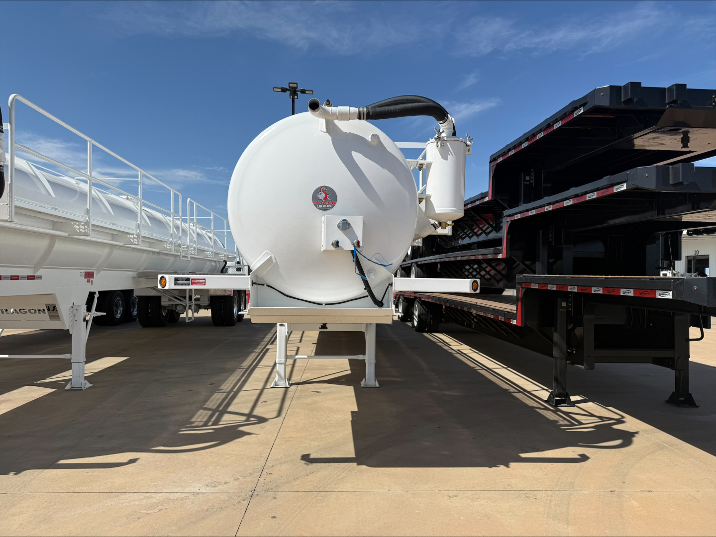 Front view of a white circular vacuum tanker trailer mounted on a stand with hoses and control valve, parked between two other trailers.