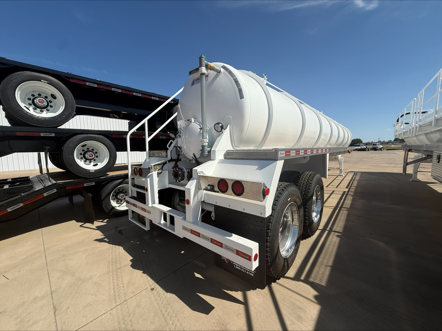 Vacuum trailer showing rear view with white cylindrical tank on dual-axle chassis and rear bumper, hoses and valves visible.