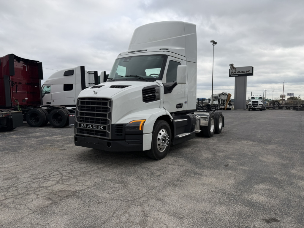 Mack AN64T semi-truck in a dealership lot, white cab, high sleeper, front grille with Mack emblem, daytime running lights, three-axle configuration with rear tires.