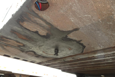 Trailer underside showing a circular area with concrete or seal around a protruding bolt, rusty metal surfaces and support rails.