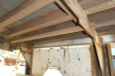 Trailer undercarriage showing riveted steel frame and rusted support beams with a white metal panel and hanging wire.