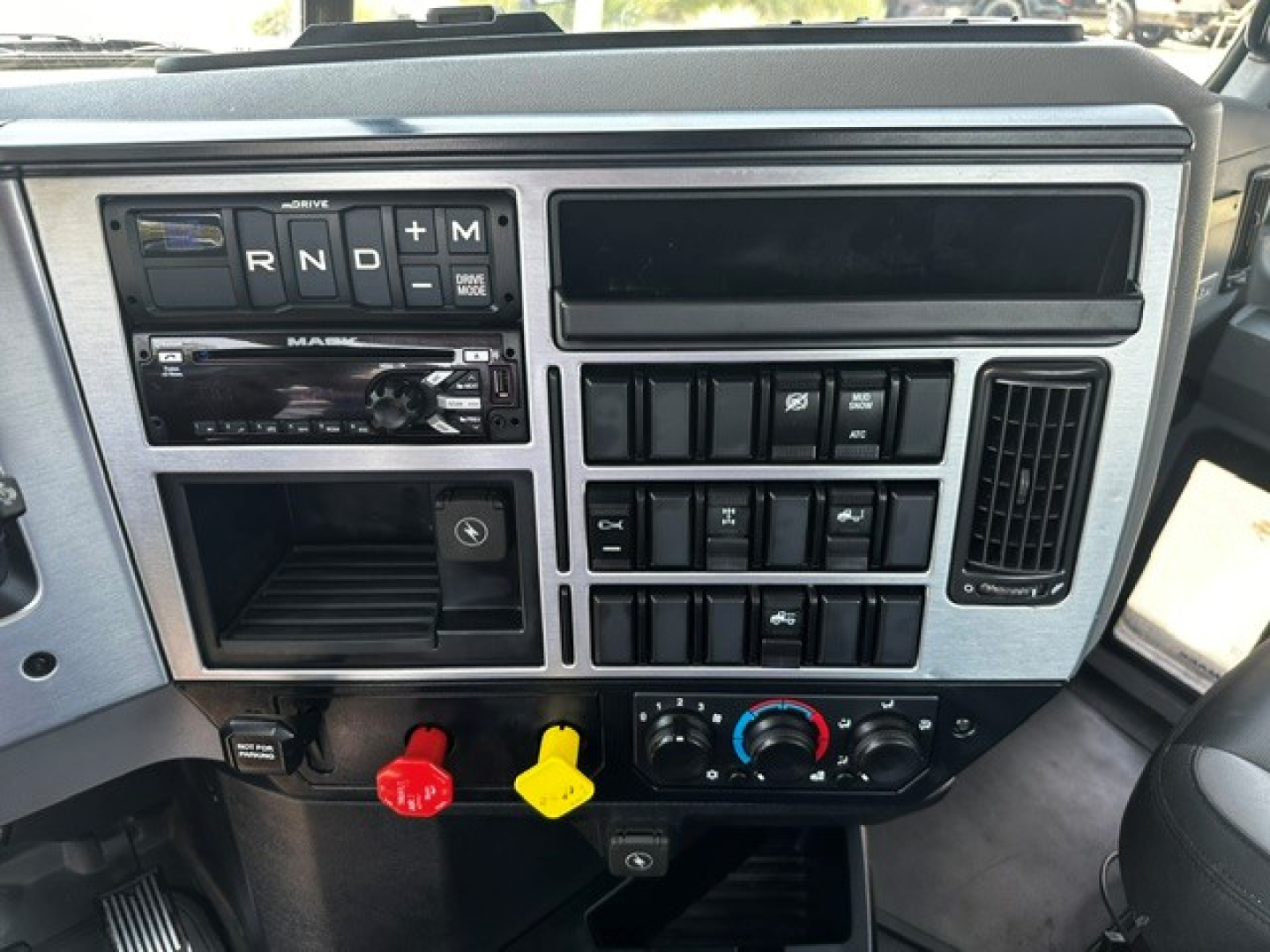 Dashboard of 2021 Mack ANTHEM showing gear selector R N D and M, climate controls, auxiliary switches, radio, and two colored knobs (red and yellow) in center console.