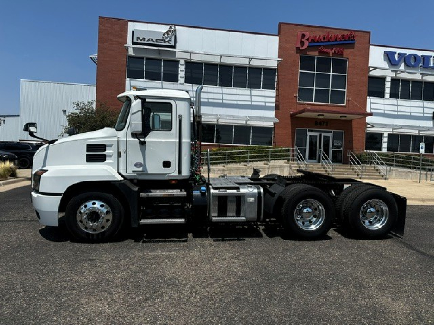 Tractor truck side view showing Mack ANTHEM chassis with cab and rear dual axle, fuel tanks, and flatbed/loafer in front of a dealership.