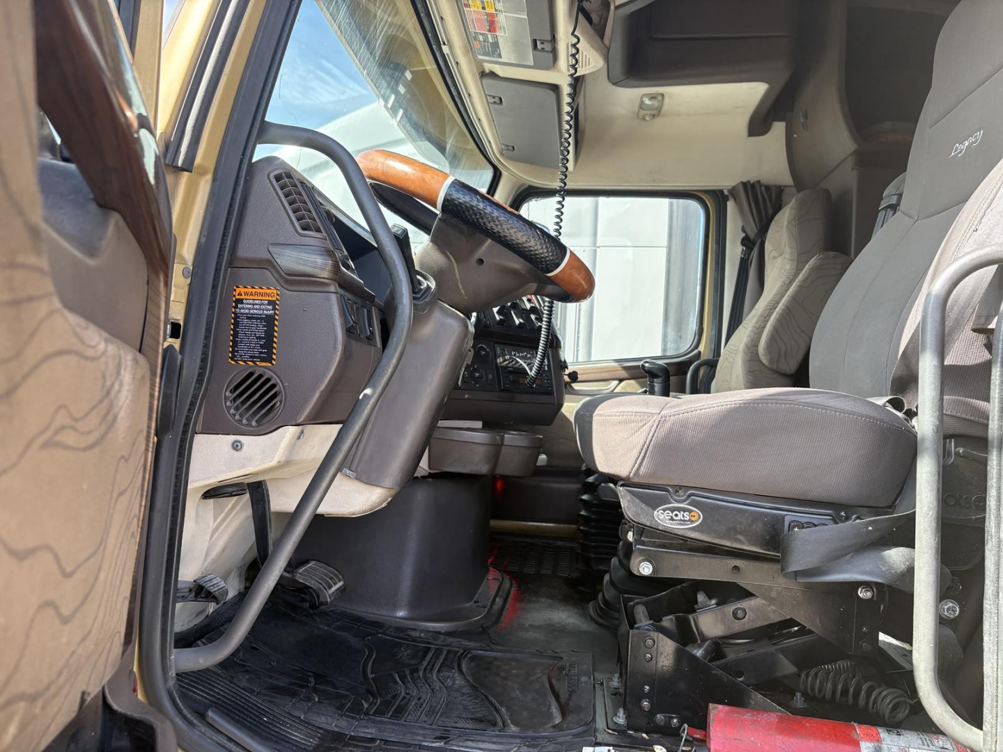 Volvo 770 sleeper cab interior showing steering wheel, dashboard, and driver seat with grey fabric; view from open door providing dash, pedals, and passenger side.
