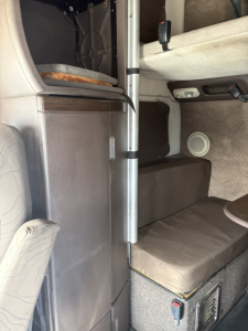 Truck cab interior showing sleeper bed area with overhead shelf and window, seating area, and control panel.