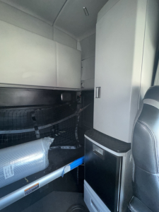 Truck cab interior showing sleeper area with storage cabinet and a mounted vertical cabinet, seat visible at right, various cables and a rolled item in the lower left.