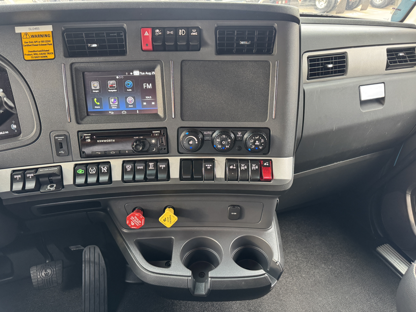 Kenworth T680 dash area showing center infotainment screen, climate controls, steering wheel left, various switches, two cup holders, red and yellow knobs, and a small storage bin.