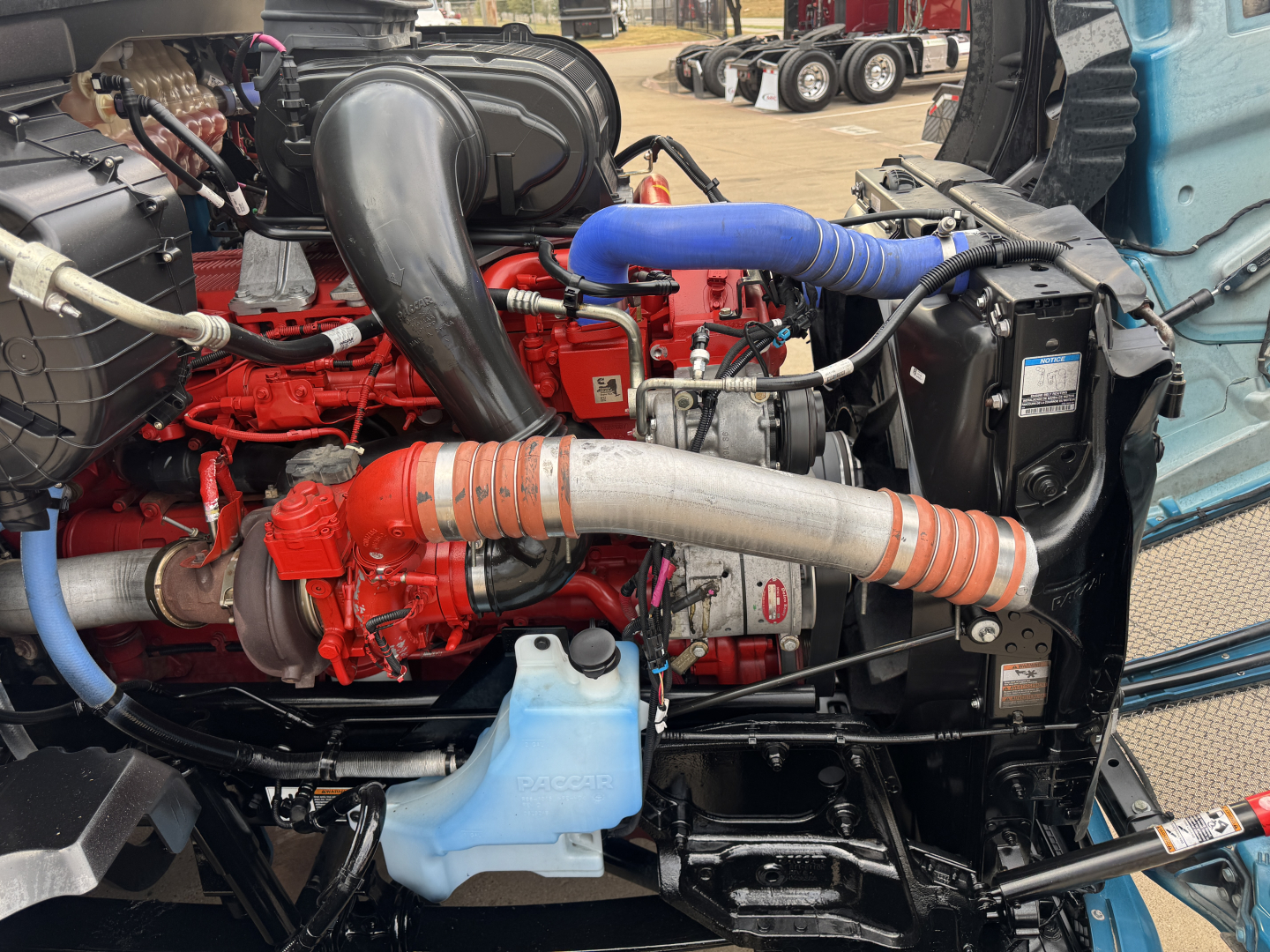 Kenworth T680 engine bay with red engine, blue hoses, coolant reservoir, and intake manifold.