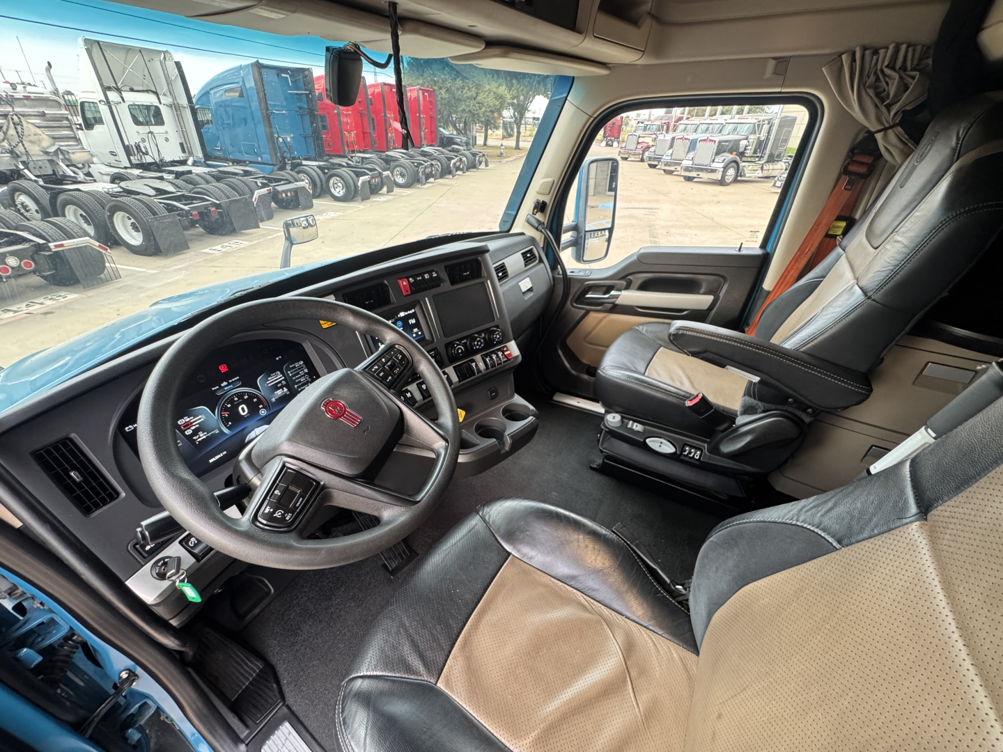 Truck cab interior of a 2024 Kenworth T680 showing steering wheel, dash, and driver's seat with beige/black upholstery.