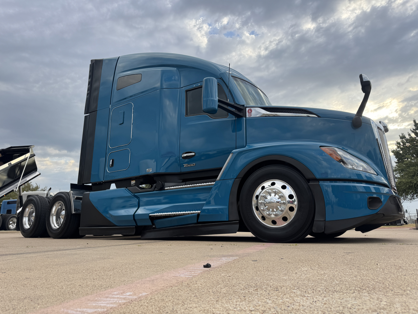 Kenworth T680 tractor cab side view showing blue exterior, sleeper cab, dual rear axles, front fender, and chrome wheels.