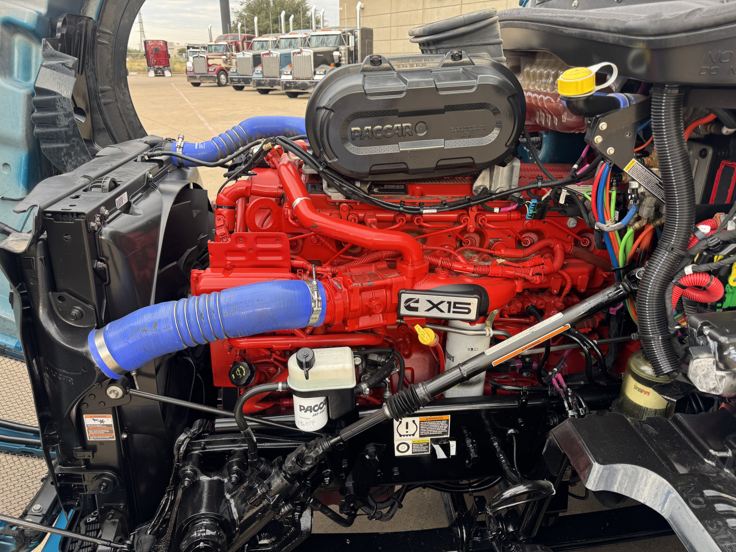 Kenworth T680 engine bay showing red X15 engine with blue coolant hoses and various wiring harnesses.