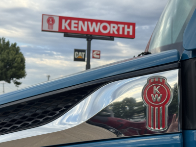 Kenworth T680 front fender badge with KW emblem and chrome grille in foreground; Kenworth sign roadside in background.