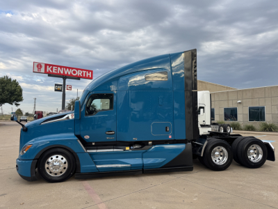 Blue Kenworth T680 tractor-trailer in a parking lot, side view showing wheel set and sleeper cab. Kenworth dealership sign visible in distance.