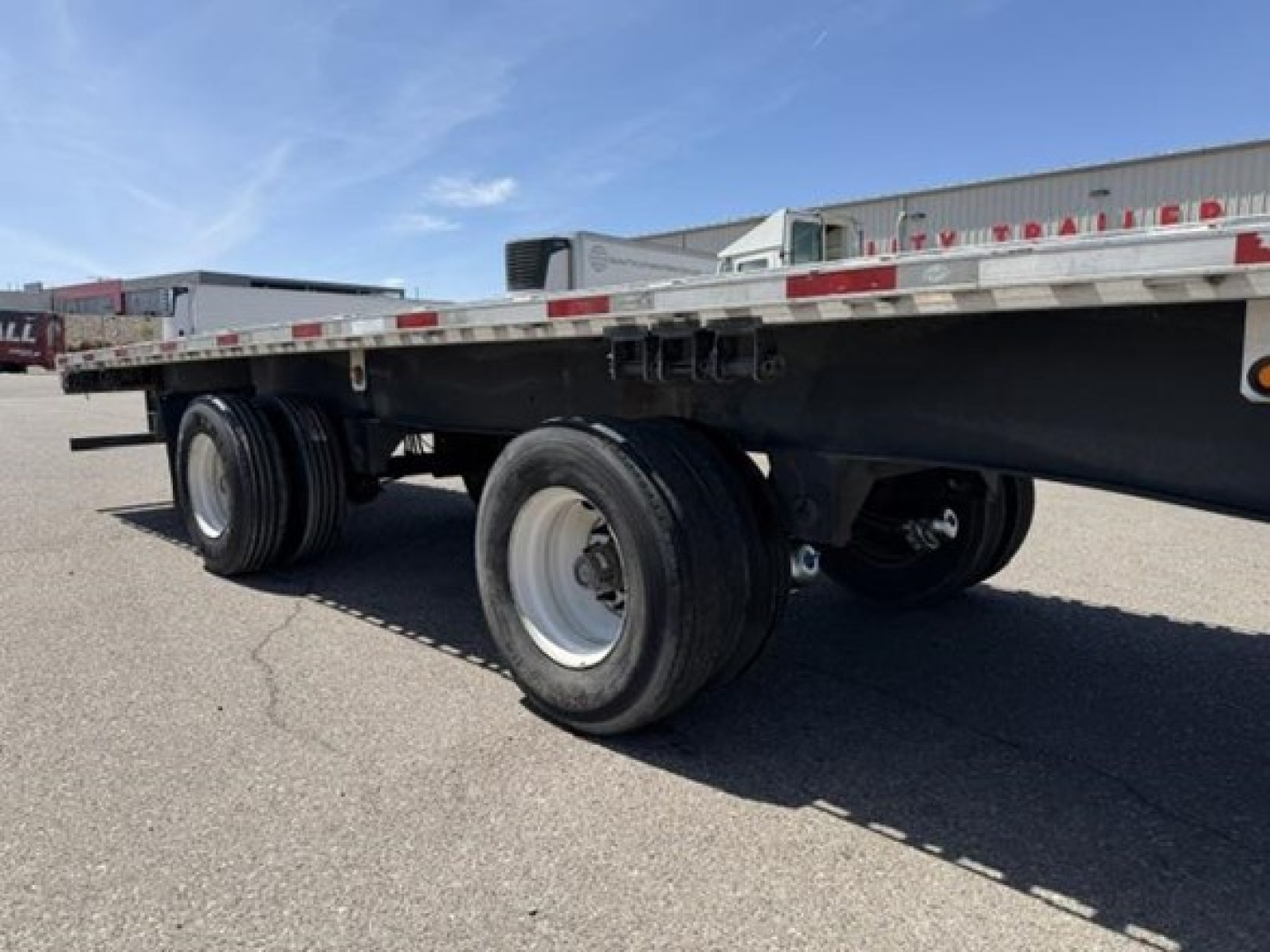 Flatbed trailer showing tandem axle wheels and thin guard rails with reflective tape along side. No legible labels visible on the frame.