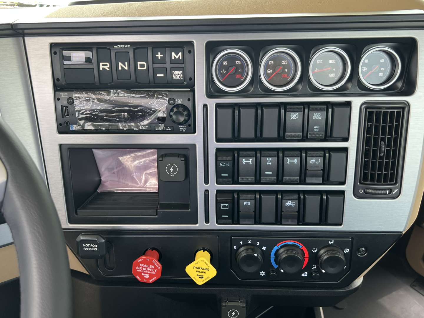 Dashboard panel of a 2027 Mack PI64T truck showing gear selector RND + M, four gauges, and various switches; red and yellow knobs, climate controls, and empty slot panels.