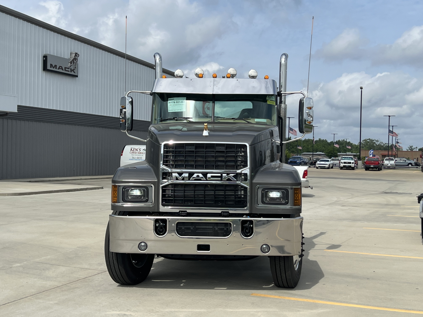 Front view of a new 2027 Mack PI64T conventional daycab truck with chrome bumper, large grille, and Mack emblem; visible windshield, headlights, and hood ornament; dealership lot in background.