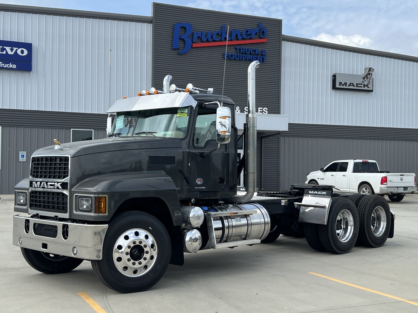 Mack PI64T conventional daycab tractor with dual rear axles and chrome fuel tanks parked in front of a Mack dealership.