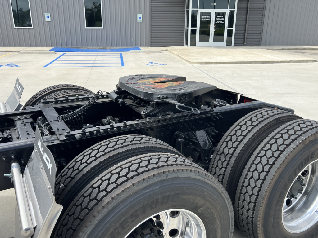 Mack PI64T truck chassis shown from rear-left side with dual rear axles and tires, fuel cap area, and vertical cab behind in a dealership lot.