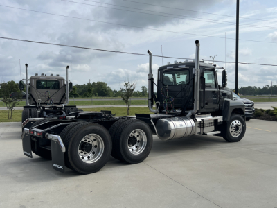 Two black Mack PI64T trucks with tandem rear axles parked in a lot, cab with chrome exhaust stacks, large fuel tanks, and dual rear wheels.