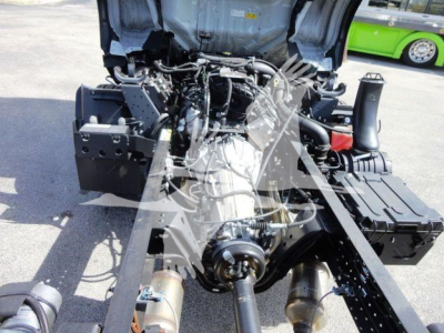 Isuzu NPR-HD engine bay view showing engine and surrounding components under open hood; visible belts, hoses, and air intake; chassis rails and brackets in the foreground.
