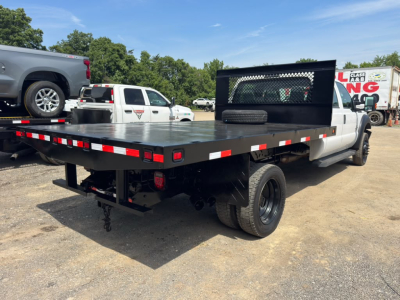 Flatbed tow truck rear and side view showing large flatbed with reflective striping, black headache rack, dual rear wheels, and hitch components visible.