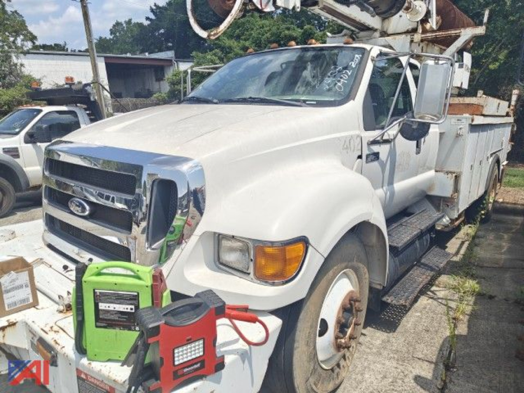 Ford F-750 Super Duty bucket truck with boom equipment and utility body, front view, white cab, green portable generator and red jumper near bumper.