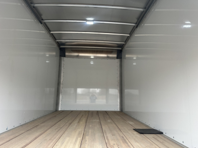 Truck trailer interior empty cargo area with wooden floor and white walls, rear doors closed, faint reflection of photographer.