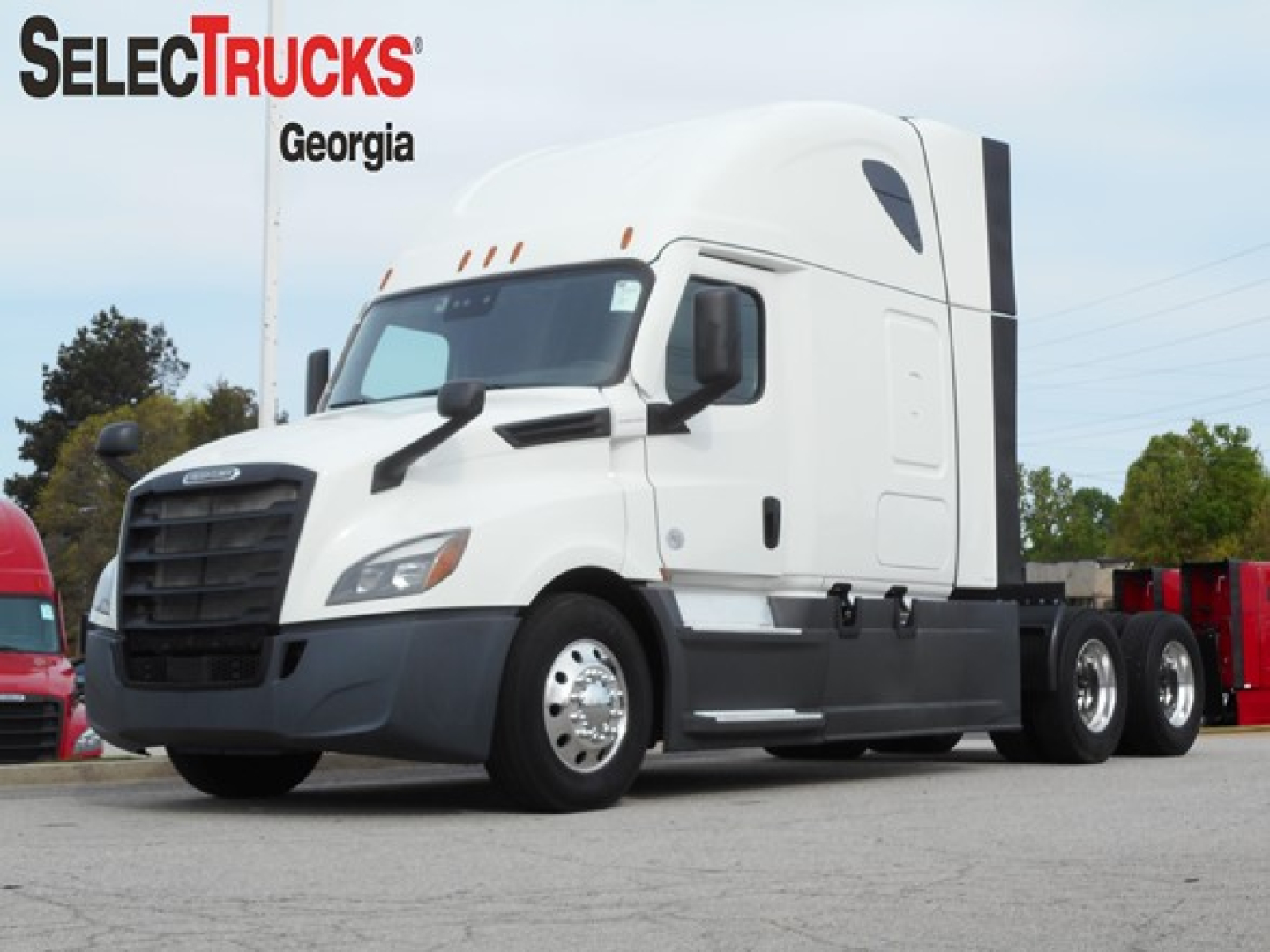 Freightliner Cascadia 126 sleeper cab truck from the front-left view. White cab with black bumper and wheels, storage compartments along side, forest green trees in background.