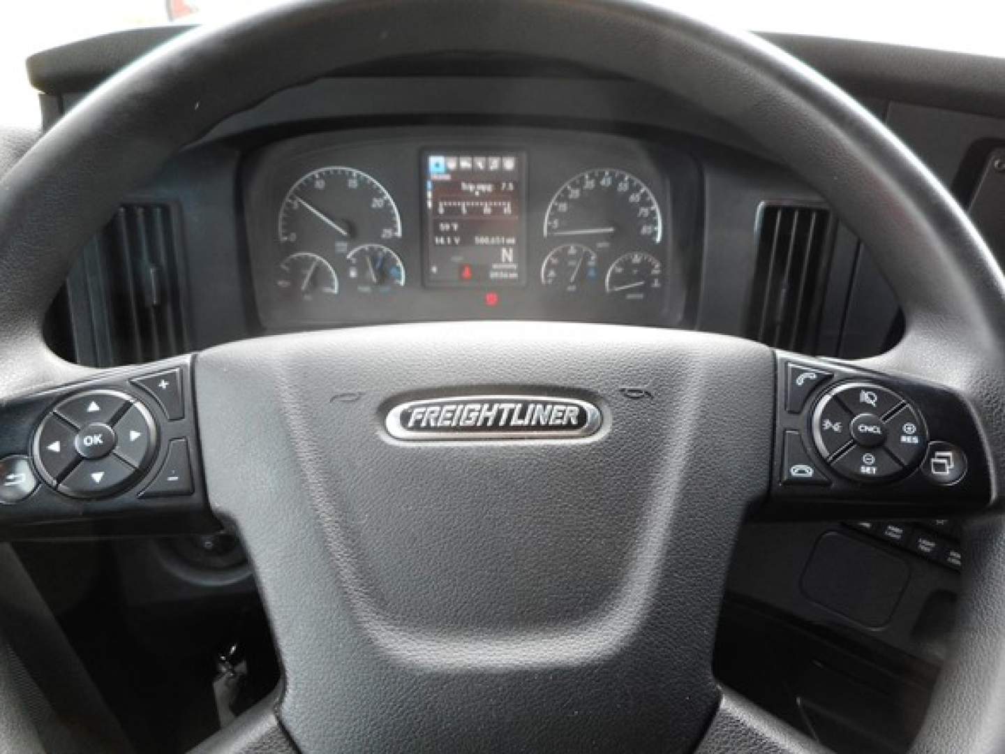 Steering wheel of a Freightliner truck with dashboard gauges visible, center screen and control buttons on both sides.
