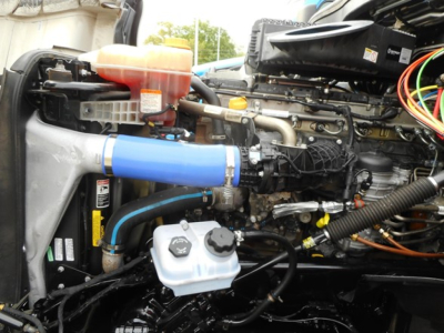 Front engine bay of a Freightliner Cascadia showing coolant reservoir, blue intercooler pipe, black hoses, and various engine components.