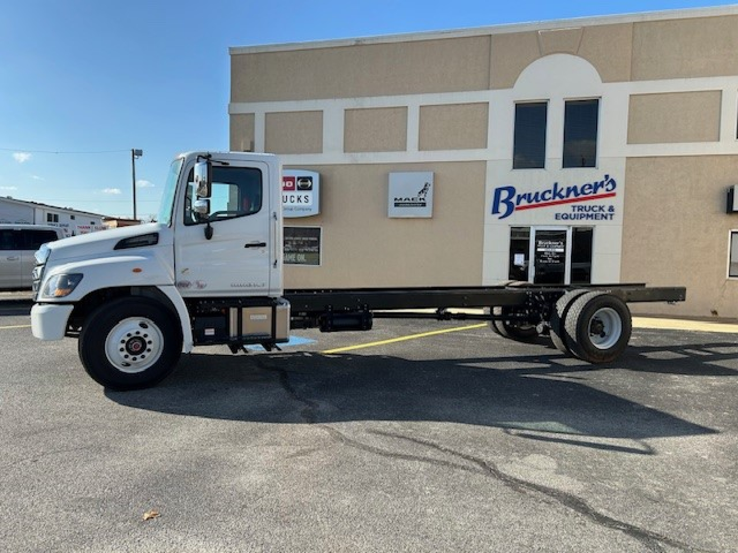 Truck chassis in front of a dealership showing a white cab and long black flatbed chassis with no cargo, 2025 Hino L7, side view, stock sign in background