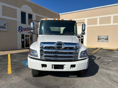 Front view of a white 2025 Hino L7 cab and chassis truck parked in a lot; grille with Hino emblem, bumper with openings, large windshield, side mirrors; building with Brücker sign in background.