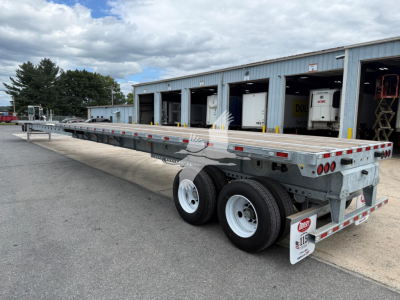 Flatbed trailer with extendable centerbeam showing diamond plate deck and dual rear axles in a yard.