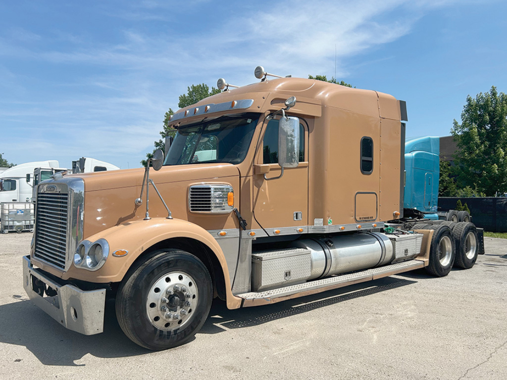Freightliner Coronado heavy-duty truck side view showing tan sleeper cab and fuel tanks, with a blue trailer attached, model details not legible.