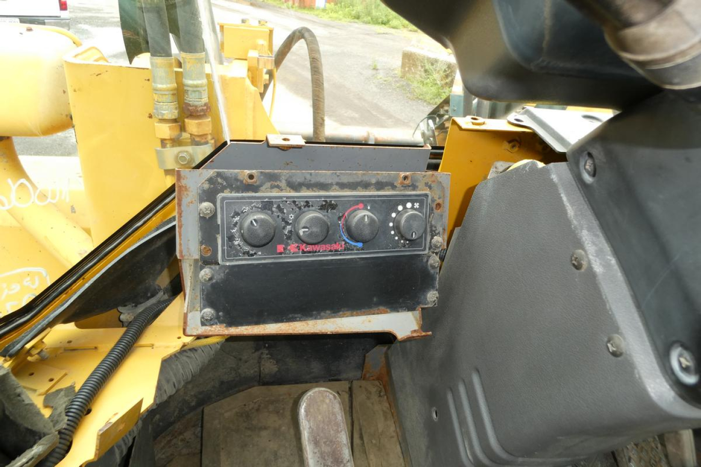 Equipment cockpit of a Kawasaki 65TMV wheel loader showing control panel with three knobs and gauges, surrounding yellow machinery and hydraulic hoses.