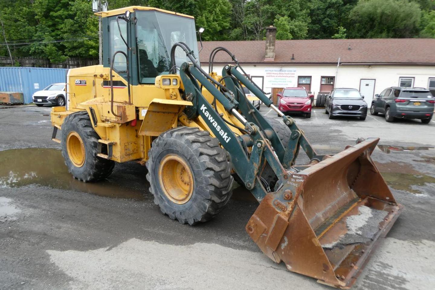 Front-left view of a yellow Kawasaki 65TMV wheel loader with a green arm and a brown dirt bucket, hydraulic lines visible, in a lot.