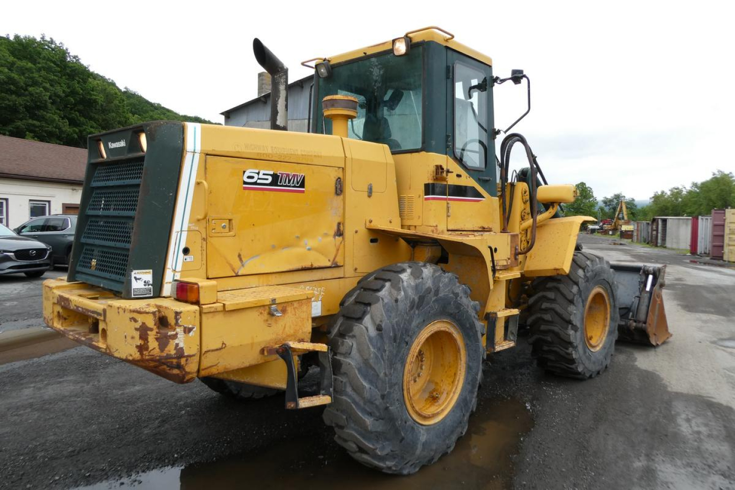 Front-left view of a yellow Kawasaki 65TMV wheel loader with large tires and a cab, showing model badge 65TMV on the side and rear.