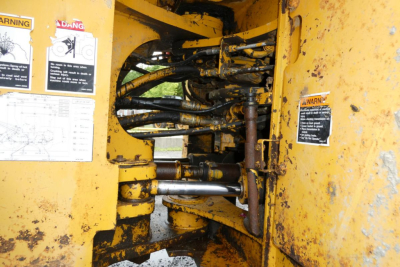 Machine hydraulic lines and controls inside the operator area of a yellow Kawasaki 65TMV wheel loader showing hydraulic hoses, levers, and warning labels.