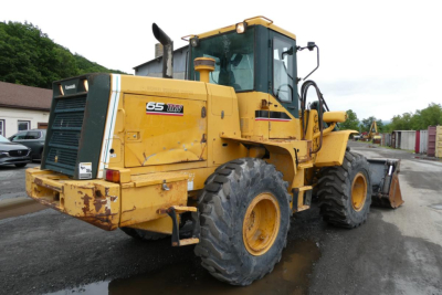 Front-left view of a yellow Kawasaki 65TMV wheel loader with large tires and a cab, showing model badge 65TMV on the side and rear.
