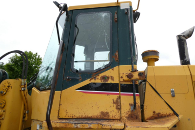 Wheel loader cabin profile with cracked windshield and rusted body panel, yellow-green paint, side window and controls visible.