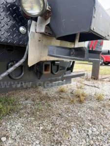 Truck rear showing plow equipment mount and bumper with metal brackets and wiring; rugged steel diamond-plate step near tail; gravel ground.