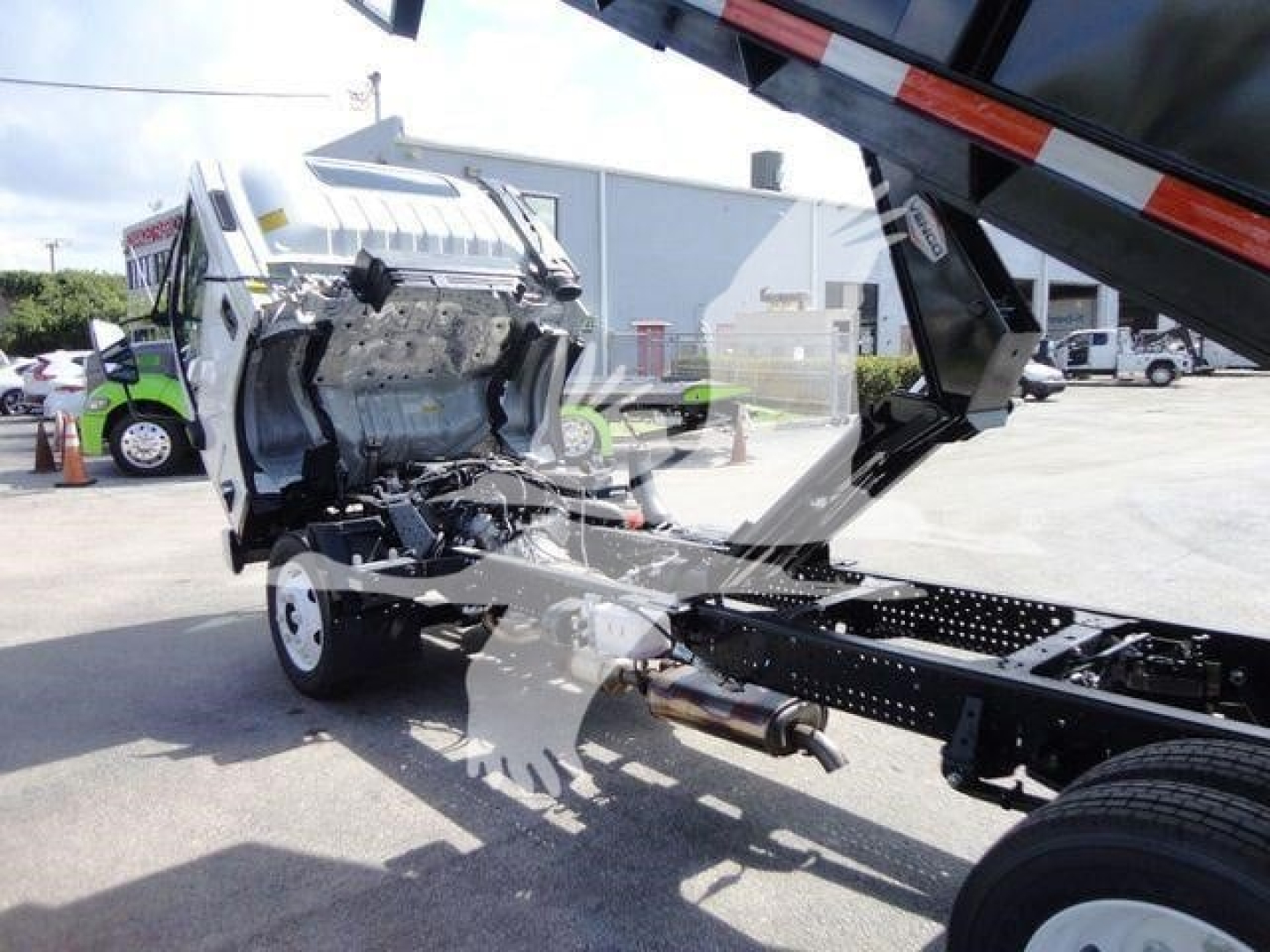 Isuzu NPR HD truck chassis with cab tilted showing engine bay area and frame; front section of cab removed; white truck, black chassis, warehouse background.