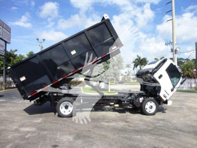 Isuzu NPR-HD dump truck with raised dump bed in service position, front cab tilted slightly, black dump body, white cab, blue sky with clouds.