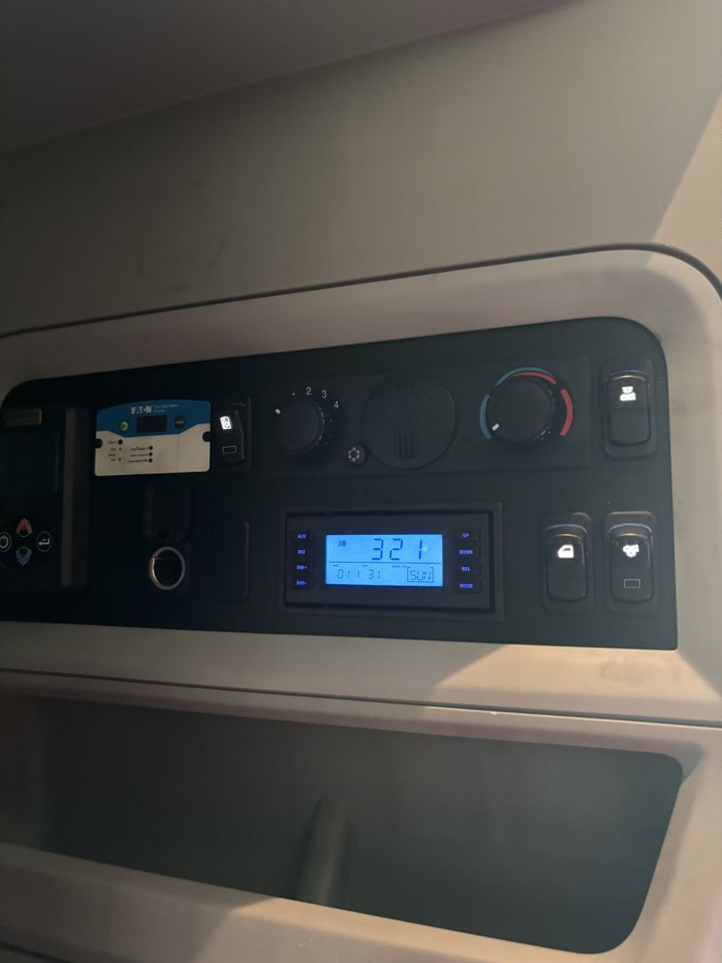Truck cab dash showing climate controls, digital display, and switches. A card and water bottle holder visible; blue-lit display reads 12:37.